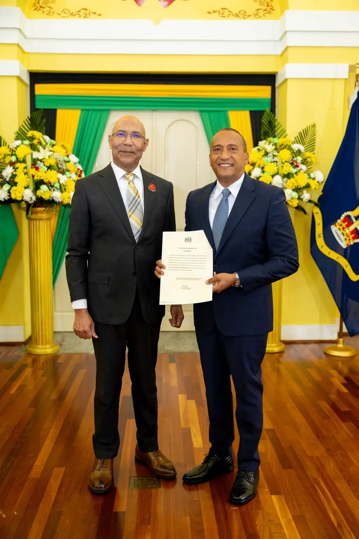 Governor General Sir Patrick Allen, on the left, and Bernard Henry, on the right, stand together at King's House holding Mr. Henry's Instrument of Appointment as Custos Rotulorum, in front of yellow, green and black drapes and yellow flowers