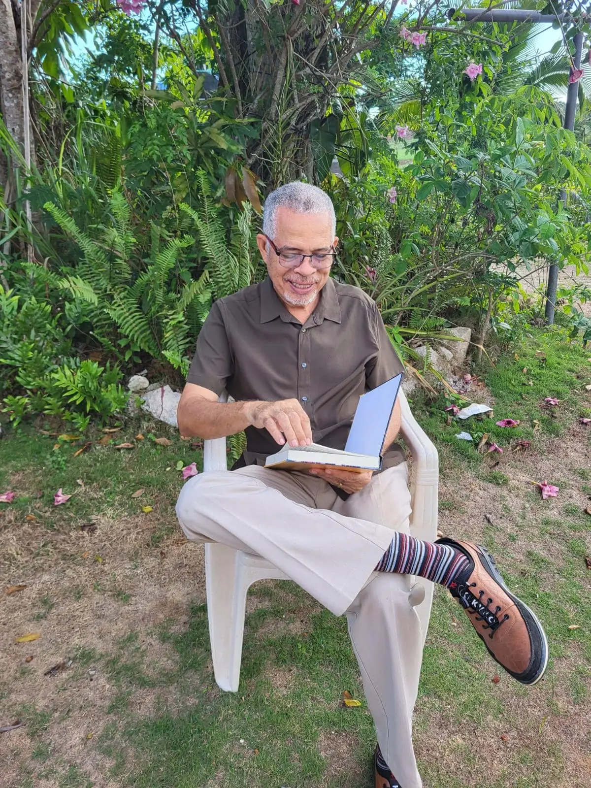 Earl Moxam, smiling, sits on a white chair in a garden and turns the pages of his book