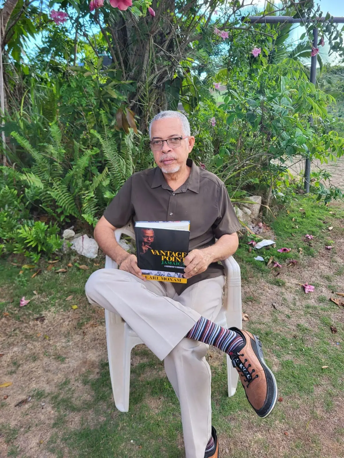 Earl Moxam, in glasses, a brown shirt and striped socks, sits cross-legged on a white plastic chair on the grass, holding his book Vantage Point Jamaica: A Reporter’s Chronicle, with ferns and a flowering tree behind him
