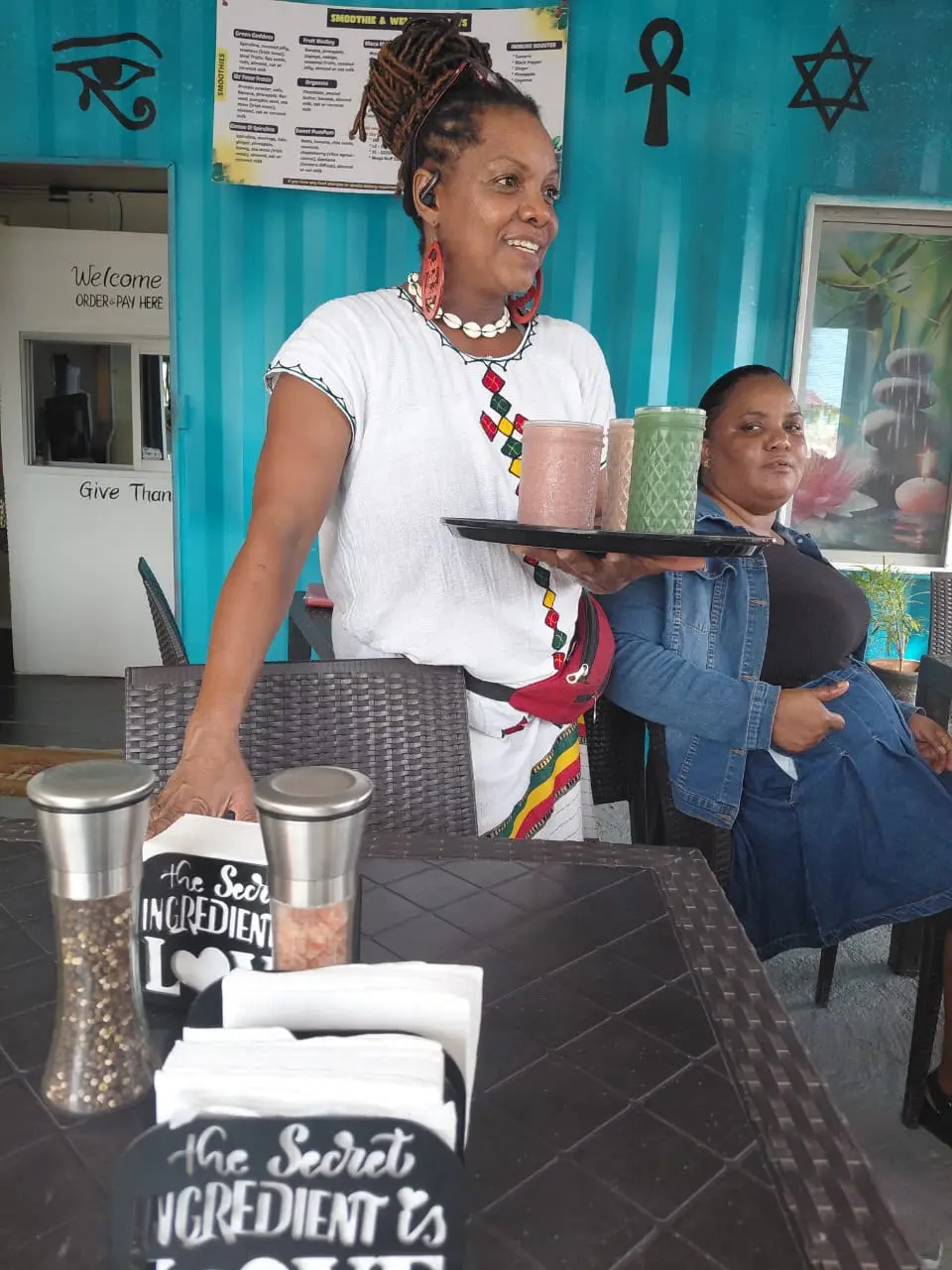 Empress Thandi Wise smiling as she carries a tray of three smoothies through the café, with the smoothie menu on the teal wall behind her