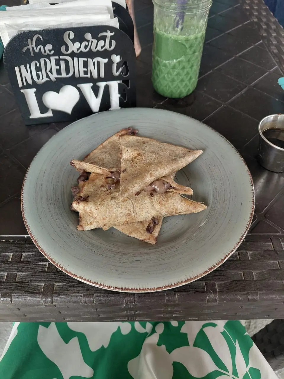 A quesadilla cut into triangles on a grey plate, with a green juice and a napkin holder reading “The secret ingredient is love”