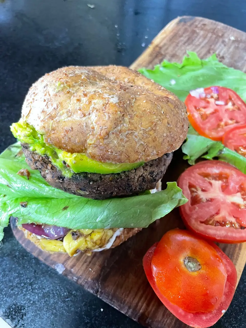 A veggie burger on a wholegrain bun with avocado, lettuce and red onion, with sliced tomatoes on a wooden board